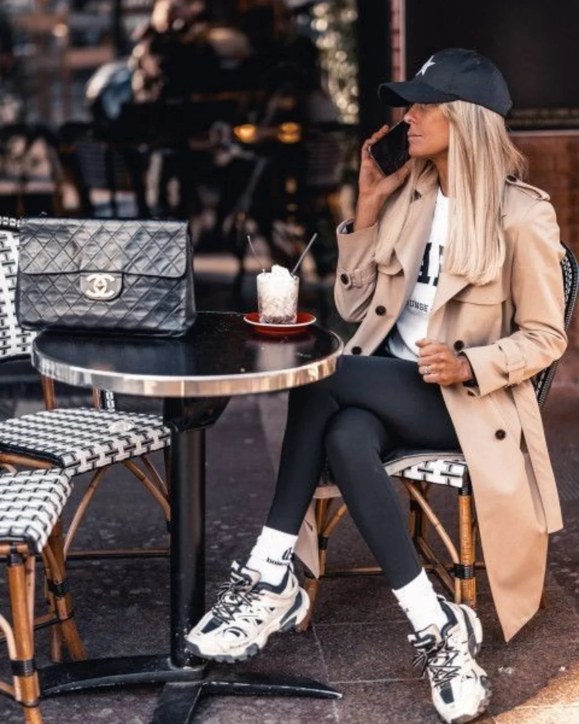 A woman in a tan trench coat and black leggings wearing chunky white sneakers while sitting at a cafe.