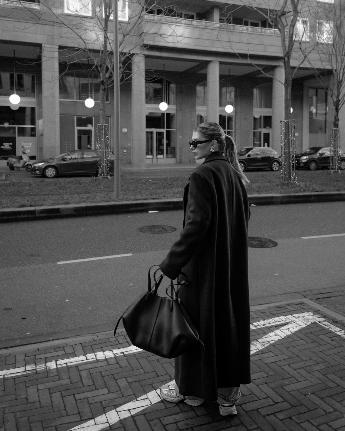 A woman wearing a long oversized black coat and sunglasses walking on a city street.