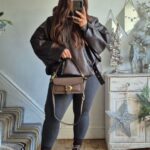 Woman in a leather jacket, grey hoodie, and leggings posing for a mirror selfie.