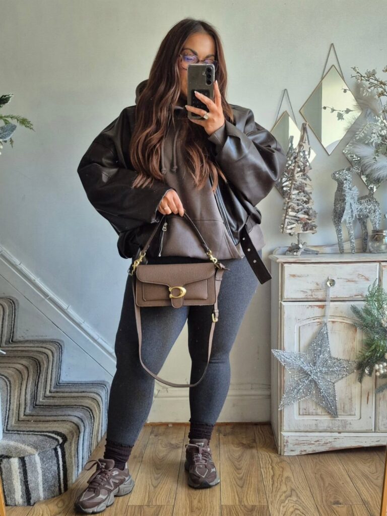 Woman in a leather jacket, grey hoodie, and leggings posing for a mirror selfie.