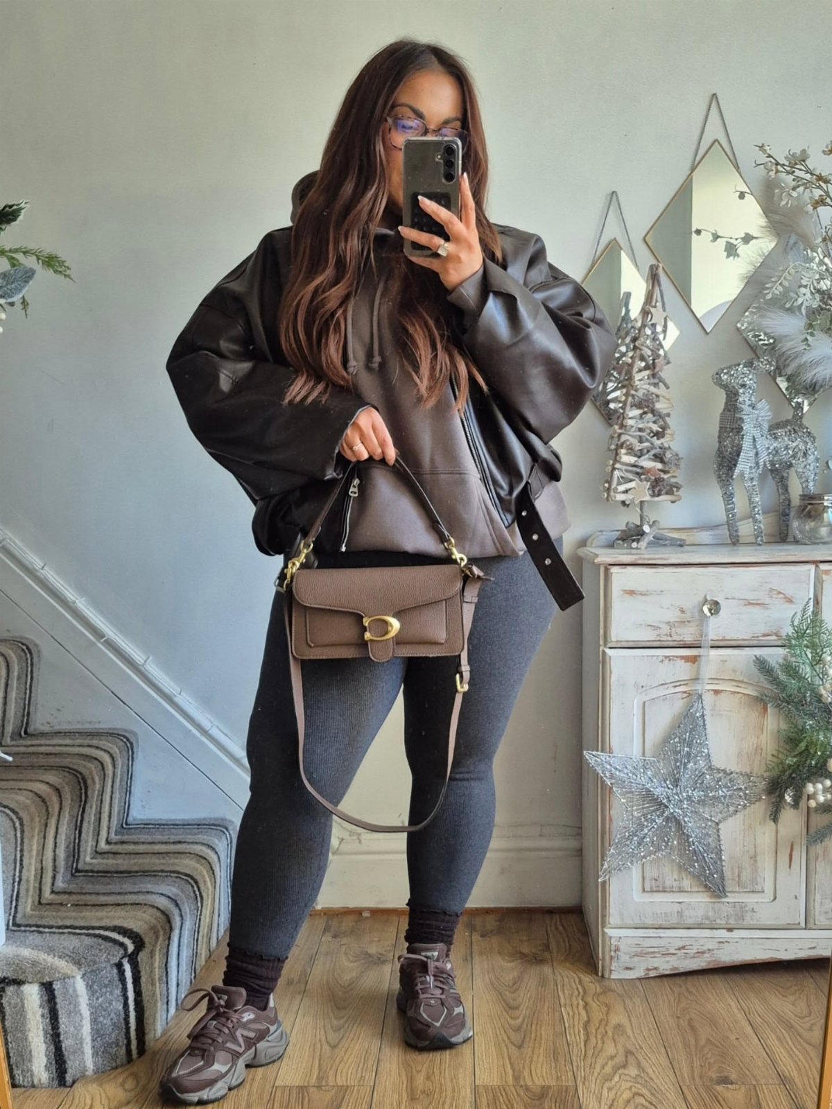 Woman in a leather jacket, grey hoodie, and leggings posing for a mirror selfie.