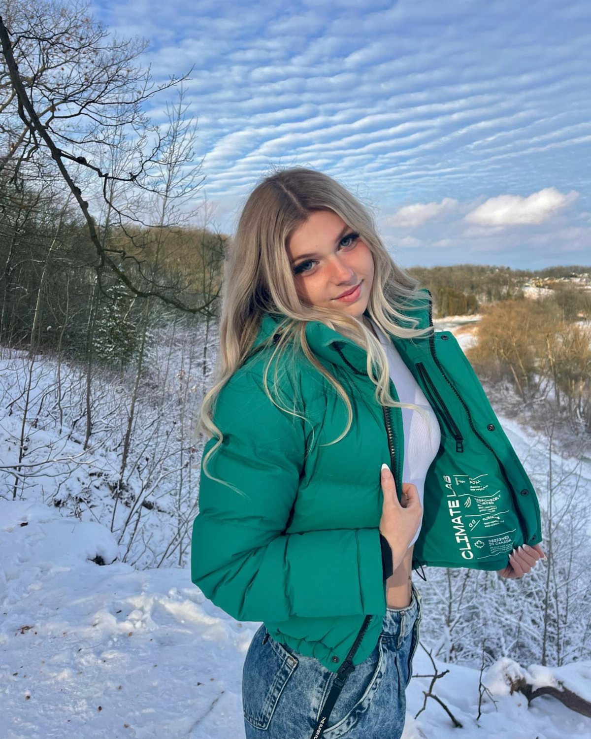 A blonde woman wearing a green puffer coat and jeans standing in a snowy landscape.