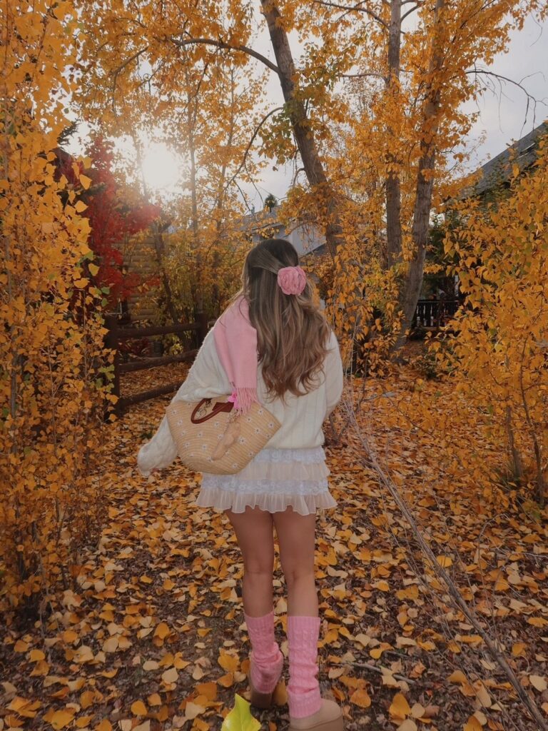 Woman in white sweater and pink accessories walking through yellow autumn leaves.