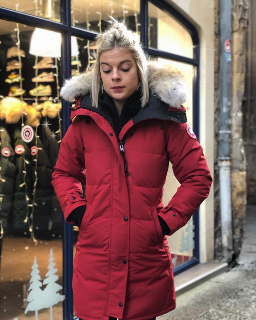 A woman wearing a long red down parka with a faux fur hood standing outdoors.