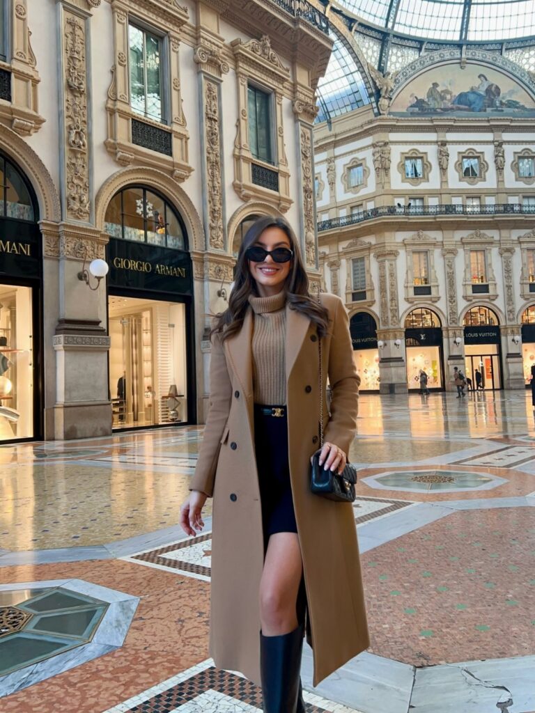 Smiling woman in a long tan wool coat and sunglasses standing in an ornate shopping arcade.
