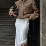 A person wearing a tan suede jacket and a white satin lace-trimmed skirt.