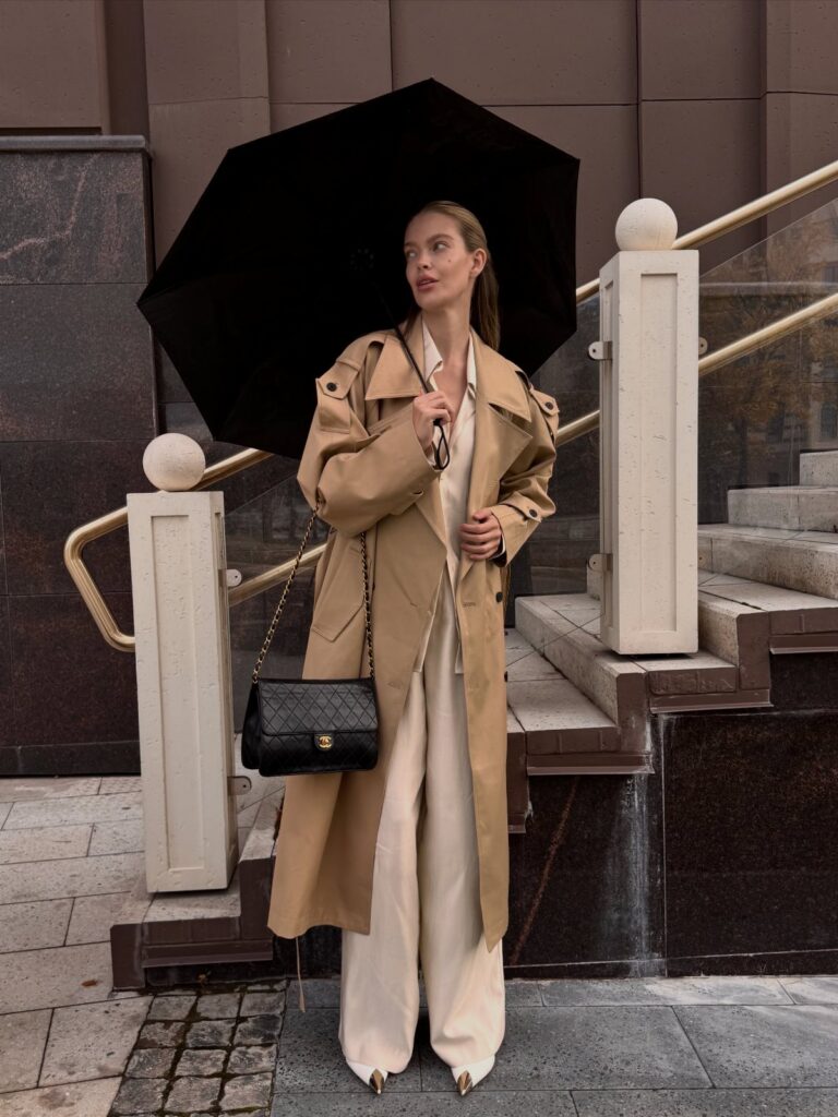 Woman in a tan trench coat and white outfit holding a black umbrella outdoors.