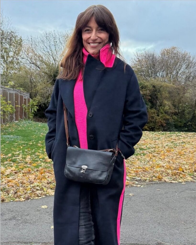 A woman smiling while wearing a bright pink turtleneck and a long black coat outdoors.