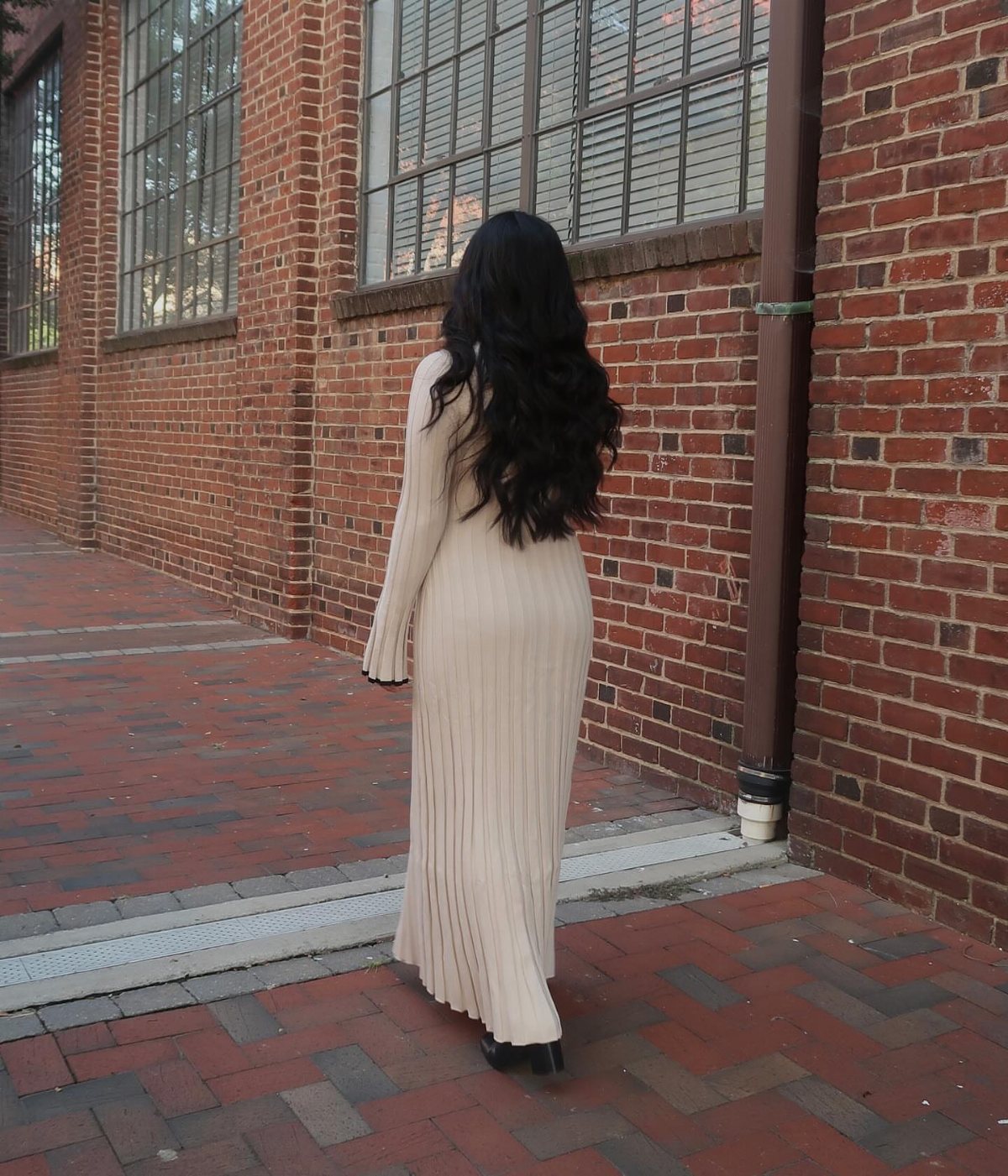 Woman in a long cream ribbed sweater dress standing against a brick wall.