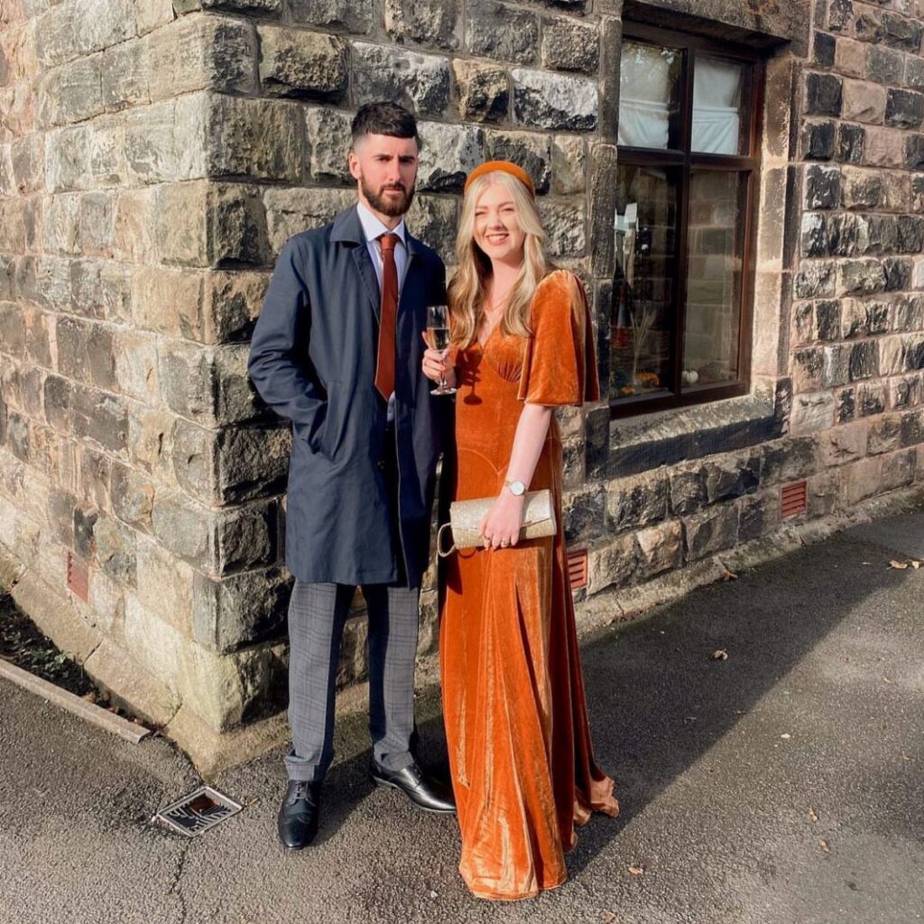 A man in a navy coat and a woman in an orange velvet dress posing outdoors.