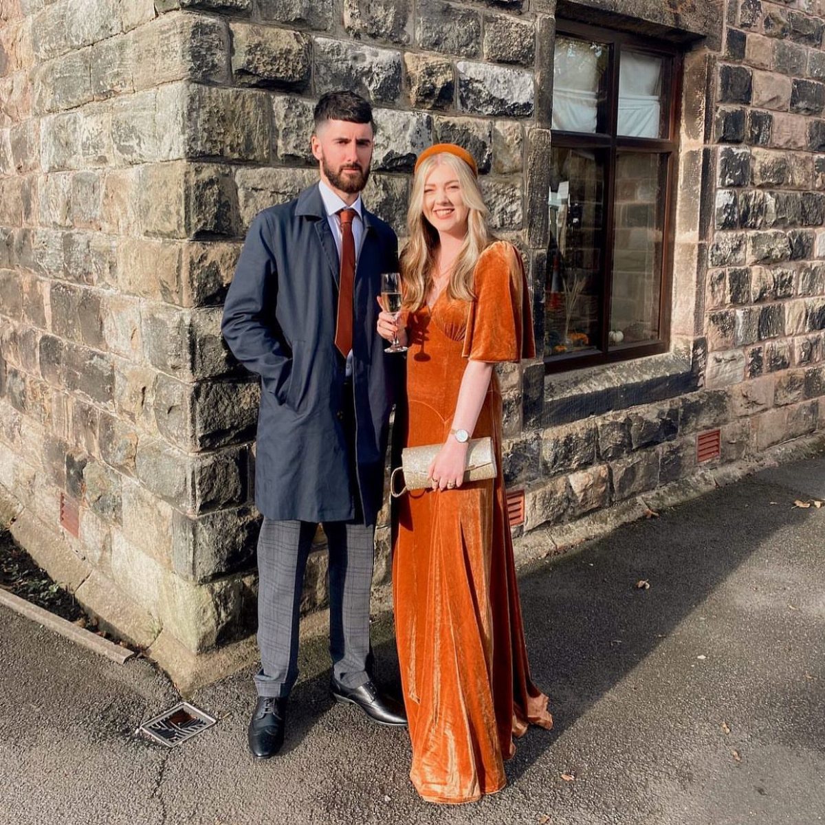 A man in a navy coat and a woman in an orange velvet dress posing outdoors.