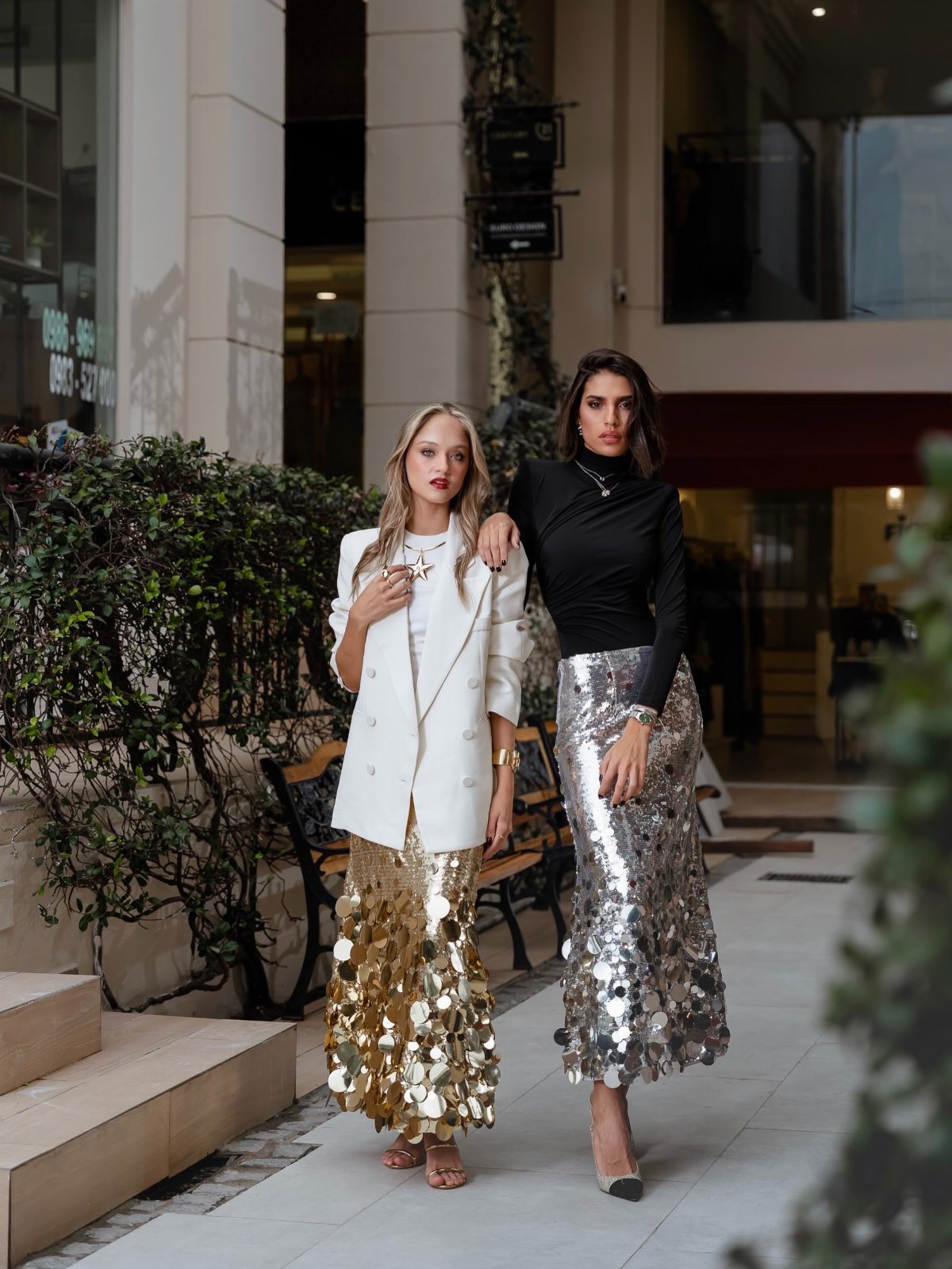 Two women posing in stylish outfits featuring gold and silver sequined metallic skirts.