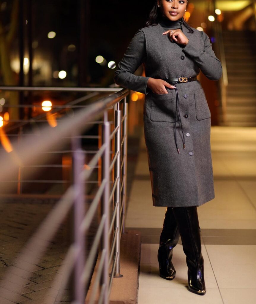 A woman wearing a long grey wool coat dress and black boots posing by a railing.