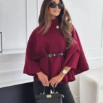 A woman posing in a burgundy cape coat cinched with a black belt and holding a handbag.