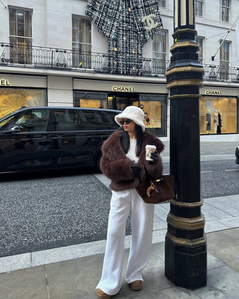 Woman in a brown faux fur coat and white trousers standing on a city street.