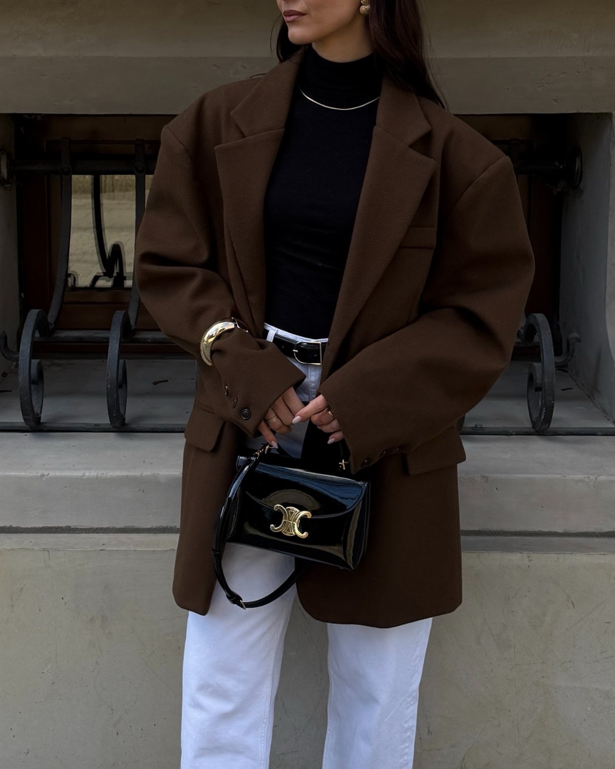 A person wearing an oversized brown blazer, black turtleneck, and white pants holding a black handbag.