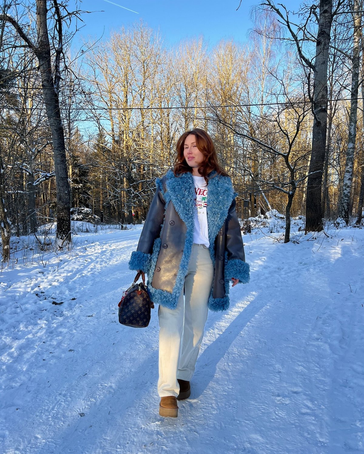 A woman in a blue fur-lined coat and white pants walking through a snowy forest.