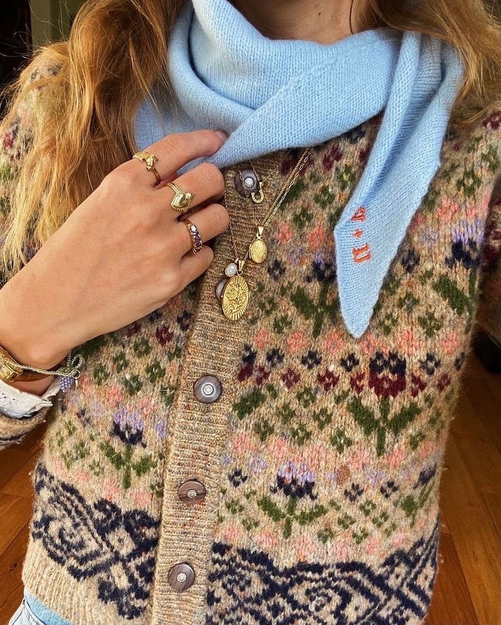 A person wearing a patterned Fair Isle cardigan with a light blue scarf and gold jewelry.