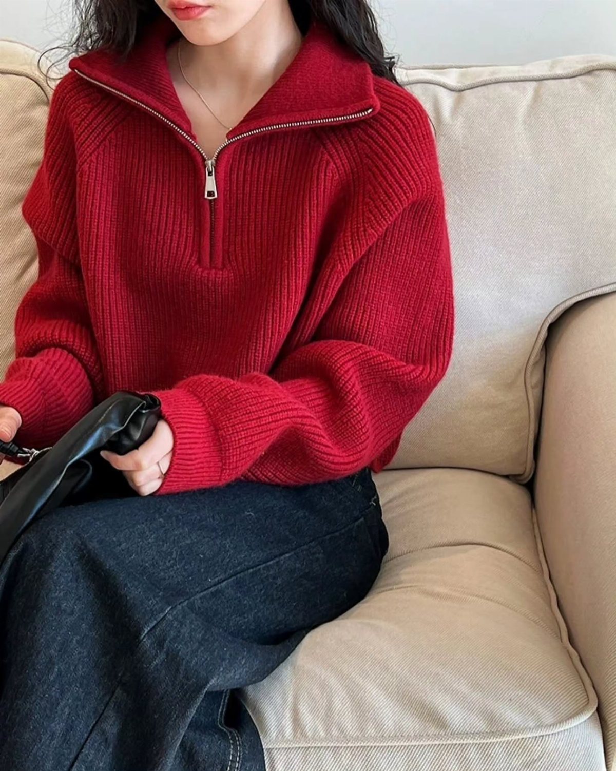 A person wearing a red ribbed quarter-zip sweater and dark jeans sitting on a beige couch.