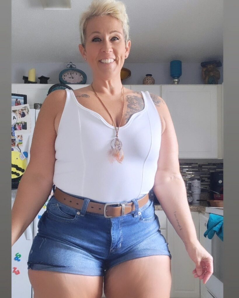 Smiling woman with short blonde hair wearing a white tank top and denim shorts.