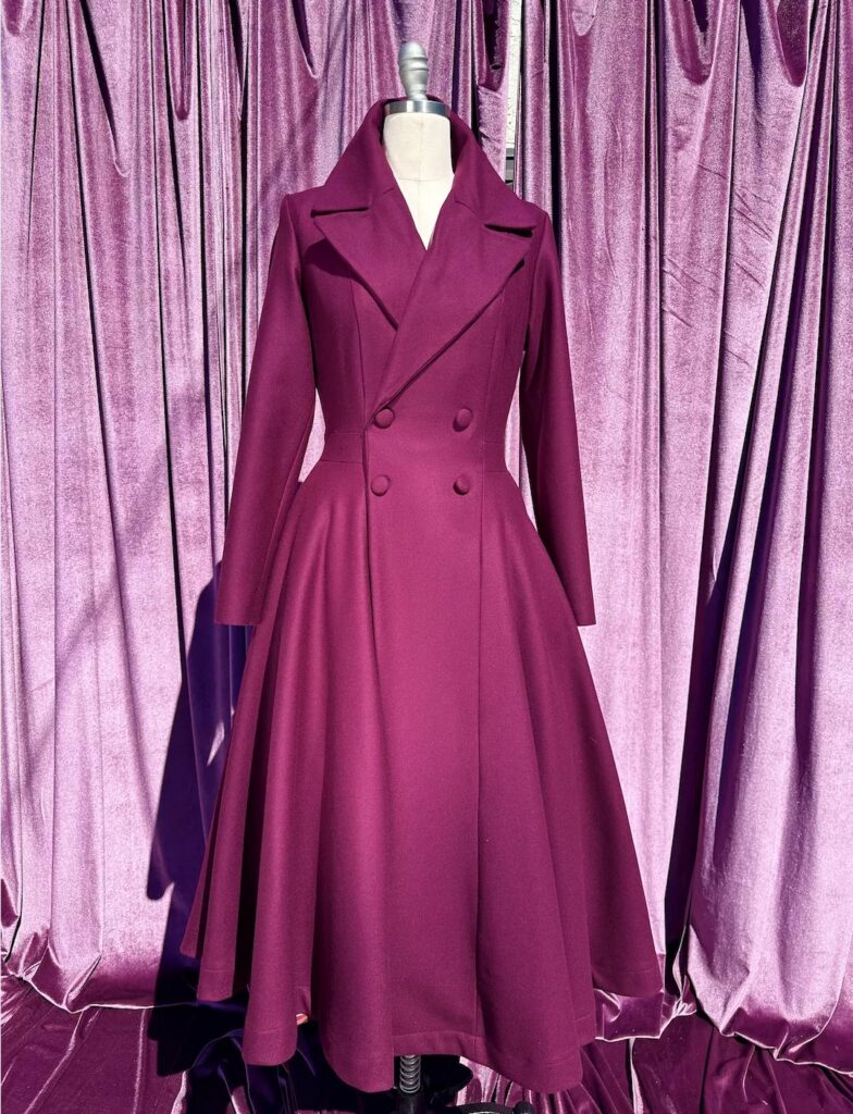 Magenta flared double-breasted coat with wide lapels displayed on a mannequin against purple curtains.