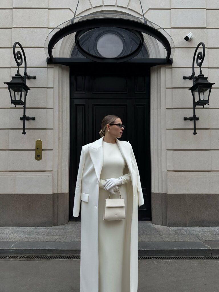 Woman in a long white wool coat and matching maxi dress standing before a black door.