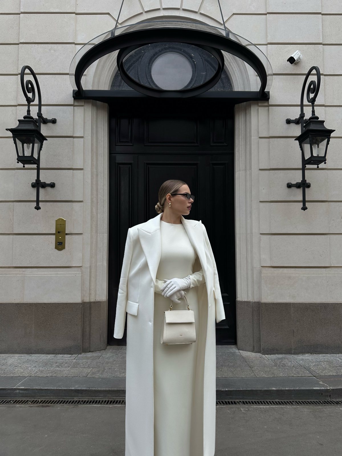 Woman in a long white wool coat and matching maxi dress standing before a black door.