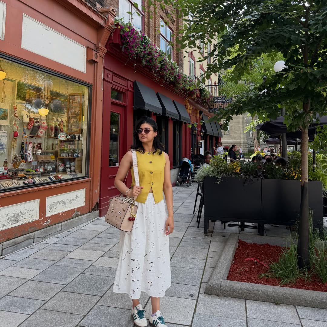 Woman in a yellow waistcoat and white skirt standing on a city sidewalk near storefronts.